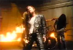 <i>Iron Maiden – Wasting Love</i>” />Epica e profanatrice, la videografia di Bayer si muove tra immagini ricorrenti che segnano inevitabilmente una poetica di grandi conflitti.<br />
 Il fuoco di <em>Wasting love</em> degli <strong>Iron Maiden</strong> è il peccato che brucia, l’estetica del metal che silenziosamente divampa. Il fuoco sta in mezzo, sta tra il vescovo che, croce alla mano,  giudica, condanna e trascina all’inferno l’uomo, e il piccolo angelo, che osserva quell’uomo, accarezzando e consolando la sua pena scritta sulla pelle.</p>
<p><strong>Il potere del fuoco</strong> è quello di mondare e di devastare: più dell’acqua, il fuoco arde ogni cosa e la purifica, segno di disperazione e di cambiamento, illumina un momento di rivelazione, dopo il quale nulla sarà più come prima.<br />
<strong>Così la croce è un segno-bersaglio:</strong> moltissimi musicisti nei video di Bayer assumono quella posizione, con le braccia allargate, diventando figure liberate e contemporaneamente in pericolo, senza possibilità di difesa. Ma la croce è anche il gesto della massima esposizione allo sguardo, della massima visibilità, un’offerta di spettacolo e morte, di sacralità sovrumana: una canzone diventata simbolo grazie ai segni del suo video è <em>Zombie</em> dei <strong>Cranberries</strong>, dove Dolores O’Riordan canta d’orovestita sotto una croce attorniata da cupidi.<br />
<strong>E gli angeli,</strong> come quello vecchio di <em>I Can’t Be With You</em>, (<strong>Cranberries</strong>), sono spesso solo donne o uomini che non portano salvezza, ma vagano senza volare, cercando un luogo migliore (forse la più bella creatura alata di Bayer è la piccola ape di <em>No rain</em> dei <strong>Blind Melon</strong>).</p>
<p>Così l’incontro con il <strong>Marilyn Manson</strong> di <em>  Disposable teens original</em> e di <em>Coma white</em>, l’estetica funerea per eccellenza, dove Bayer [img4]esprime la sua visione sporca e dai colori invecchiati, insieme a una composizione patinata dell’immagine, dove le croci diventano ambiente e dove l’angelo è solamente una vecchia Marylin Monroe (<em>Coma white</em> in particolare, con la sua parata mortuaria, viene ripreso in <em>The Black Parade</em> dei <strong>My Chemical Romance</strong>,  dove la cenere si sostituisce ai coriandoli, dove le croci diventano teschi  e Marilyn Manson si trasforma nella più rassicurante e bionda  figura di Gerard Way).</p>
<p><strong>Una purezza sempre contaminata, un angelo che non vola, una  croce che non germoglia:</strong> un matrimonio senza sacralità che finisce per essere toccato dal fuoco quello nel video di <strong>John Mellencamp</strong>, <em>Your Life Is Now</em>; un fuoco tondeggiante che segna la morte per la traditrice di <em>What Goes Around Comes Around</em> di <strong>Justin Timberlake</strong>; una sacralità demoniaca quella di <em>Until it sleeps</em> dei <strong>Metallica</strong> (video ispirato ai quadri di Hieronymous Bosch), dove la croce e le immagini divine non consolano, ma ricordano all’uomo il suo peccato originale incancellabile. </p>
<p><strong>Suburbia</strong><br />
Luoghi abbandonati, stanze-rifugio lontane da ogni civiltà, ambienti senza nome, ma che parlano di solitudine e abbandono, di scarto e di una fine imminente.<br />
<strong>L’oltre cittadino è il luogo privilegiato</strong>, capannoni, fabbriche, stanze, strade sfatte, insomma, <em>periferia</em>. Un luogo che è lontano da tutto, comunque via da qui. Per gente persa, senza speranza e sola. I luoghi nella videografia di Bayer sono sempre stati <em>Boulevard of broken dreams.</em><br />
<i><TABLE align=right cellpadding=3 border=0><TR><TD align=center valign=center><img src='articoli/blink.jpg' alt='<br /><i>Blink 182 – Stay together for the kids</i><br />‘><br /><i>Blink 182 – Stay together for the kids</i><br /></TD></TR></TABLE></i><br />
Sono le storie degli ultimi, di quelli che sono rimasti lontani, tra sporcizia e spazzatura. <strong>Spazzatura che esce dalla pellicola e ci finisce sopra</strong>, in quel modo tipico di Bayer, che tratta i fotogrammi con bruciature di sigarette e graffi (un esempio per tutti <em>Stupid girl</em> dei <strong>Garbage</strong>, appunto.) Tutti luoghi dell’oltre (-città, -vita, -tempo, -realtà, -show): la casa della folle Juliette Lewis in <em>Come to my window</em> di <strong>Melissa Etheridge</strong>, le cave di <em>Bullet With Butterfly Wings</em> (<strong>Smashing Pumpkins</strong>), i paesaggi campestre/desertico in <em>Ode to my family</em> (<strong>Cranberries</strong>), di <em>Home</em> di <strong>Sheryl Crow </strong>e di <em>Just Another Day</em> (<strong>John Mellencamp</strong>), l’improbabile piattaforma sul nulla di <em>Dig in</em> (<strong>Lanny Kravitz</strong>), la casa in demolizione di <em>Stay together for the kids</em> dei turbati <strong>Blink 182</strong>…<br />
<strong>E quando La Città c’è, è la più grande e improbabile, è una New York dell’impossibile</strong>, dai grattaceli di <em>I saw the light</em> (<strong>The The</strong>) alle mostruose identità di <em>Anybody seen my baby</em> dei <strong>Rolling Stones</strong>, dalla decadenza di <em>Angels</em> di <strong>Robbie Williams</strong> (ma potrebbe essere una qualsiasi città malfamata), ai marciapiedi di <em>Heart In A Cage</em>, degli <strong>Strokes</strong>.</p>
<p>Un percorso che ha portato alla festa malata di <em><A href=